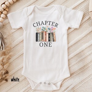 May include: White baby onesie with short sleeves. The onesie features the text "CHAPTER ONE" above a graphic of books with flowers. The word "white" is printed at the bottom left of the onesie.