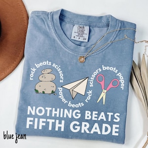 May include: Blue jean colored t-shirt with the text "NOTHING BEATS FIFTH GRADE". The shirt features a "rock, paper, scissors" design with a stack of rocks, a paper airplane, and pink scissors. A gold necklace is draped across the shirt.