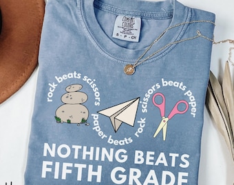 Peut inclure: Un t-shirt bleu jean avec un motif représentant un empilement de pierres, un avion en papier et des ciseaux roses. Le texte indique rock beats scissors, scissors beats paper, paper beats rock et NOTHING BEATS FIFTH GRADE en blanc.