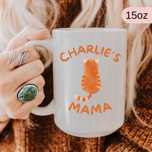 May include: White ceramic mug with the text "Charlie's Mama" in orange, featuring an orange cat illustration. The mug is 15oz in size.