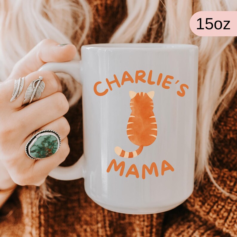 May include: White ceramic mug with the text "Charlie's Mama" in orange, featuring an orange cat illustration. The mug is 15oz in size.