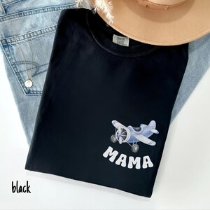 Pode incluir: T-shirt preta com um desenho de avi&atilde;o azul claro e a palavra "MAMA" em branco. A camisa est&aacute; dobrada e colocada em cima de um par de jeans azuis e um chap&eacute;u bege. A palavra "black" est&aacute; escrita no canto inferior esquerdo.