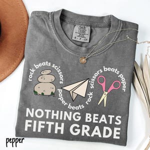 May include: A gray Comfort Colors t-shirt with a round neck, featuring a "rock, paper, scissors" design and the text "NOTHING BEATS FIFTH GRADE." The design includes a stack of stones, a paper airplane, and pink scissors.