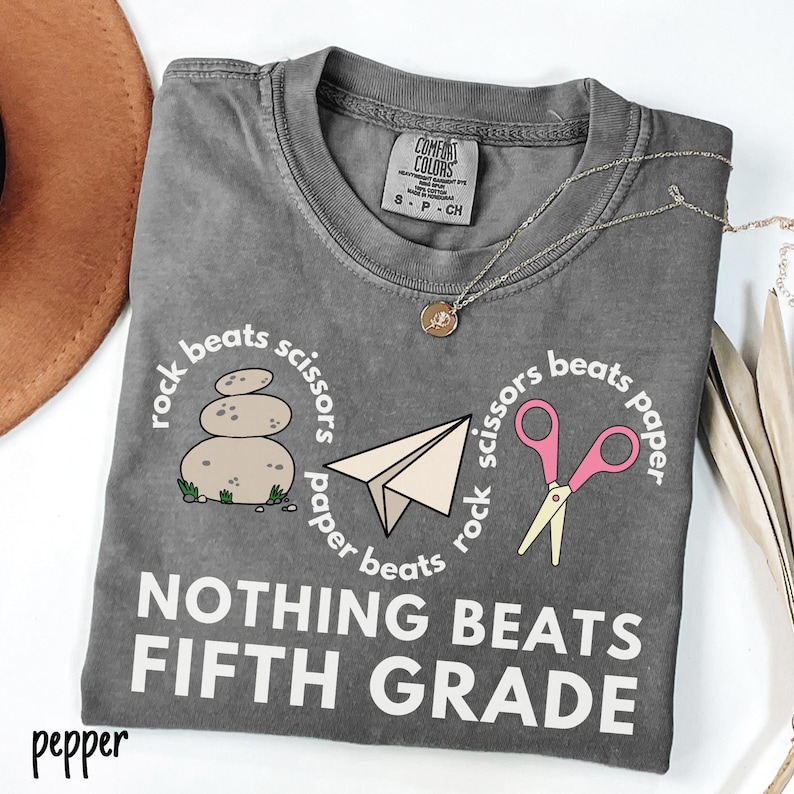 May include: A gray Comfort Colors t-shirt with a round neck, featuring a "rock, paper, scissors" design and the text "NOTHING BEATS FIFTH GRADE." The design includes a stack of stones, a paper airplane, and pink scissors.