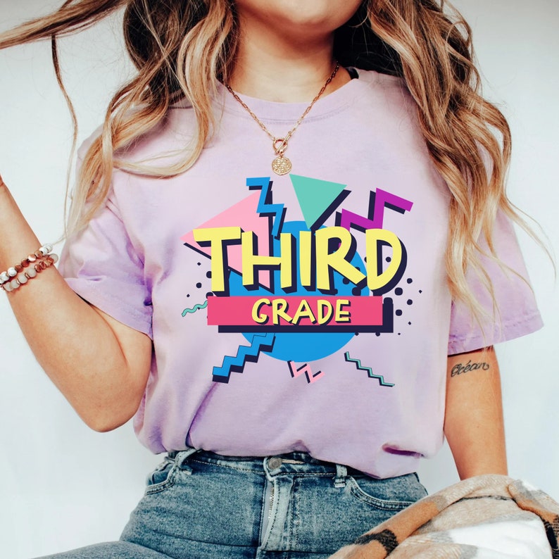 Op de afbeelding: Lichtpaars T-shirt met de tekst "THIRD GRADE" in een kleurrijk grafisch ontwerp. Het retro-ontwerp uit de jaren 80 en 90 maakt gebruik van geometrische vormen en heldere kleuren. Het T-shirt is gemaakt van zacht materiaal.