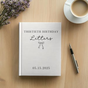 May include: A white book with the text "THIRTIETH BIRTHDAY Letters" and the date "05.15.2025" printed on the cover. A silver pen and a cup of coffee are next to the book. A bouquet of purple flowers is in the background.