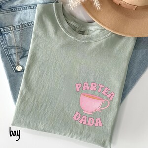 Puede incluir: Camiseta verde claro con las palabras rosas "PARTEA DADA" y un dibujo de taza de t&eacute; rosa. La camiseta est&aacute; colocada sobre unos vaqueros azules, con un collar dorado y un sombrero beige en el fondo.