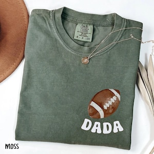 May include: Moss green t-shirt with a football graphic and the word "DADA" in white. The shirt has a crew neck and short sleeves. A silver necklace with a pendant is draped on the shirt.