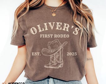 May include: Espresso-colored t-shirt with the text "OLIVER'S FIRST RODEO" above a graphic of a cowboy hat and boots. The text "EST. 2025" is below the graphic. The shirt is worn by a person.