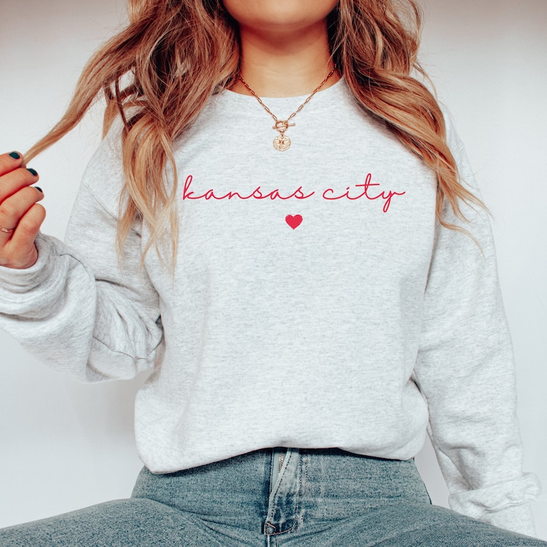 May include: A gray sweatshirt with the text "kansas city" in pink script with a small pink heart below the text.