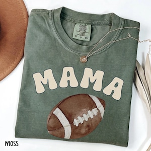 May include: A moss green t-shirt with the word "MAMA" in cream-colored, retro-style lettering and a watercolor football graphic. A gold necklace with a pendant is draped on the shirt.