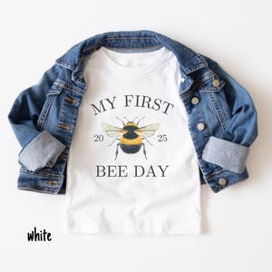 May include: White t-shirt with a black and yellow bee graphic and the text "My First Bee Day" with the numbers 20 and 25 on either side of the bee.