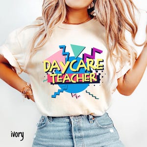 May include: Ivory-colored t-shirt with a colorful graphic design. The design features the words "DAYCARE TEACHER" in yellow, outlined in black, with a retro 90s style. The graphic includes geometric shapes in pink, blue, green, and purple.