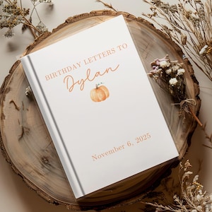 May include: A white hardcover book with the text "BIRTHDAY LETTERS TO Dylan" in orange script, a small pumpkin illustration, and the date "November 6, 2025". The book is on a wooden slice with dried flowers.