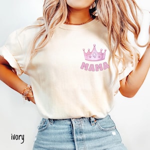 May include: Ivory-colored t-shirt featuring a pink crown design above the word "MAMA" in a bubble font. The shirt is a relaxed fit and has short sleeves. The word "ivory" is written in a script font at the bottom left.