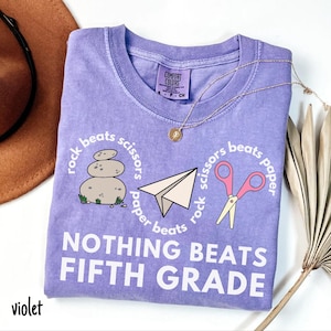 May include: A violet Comfort Colors t-shirt with a round neck and short sleeves. The shirt features a "rock, paper, scissors" design with the text "NOTHING BEATS FIFTH GRADE". A gold necklace is draped across the shirt.