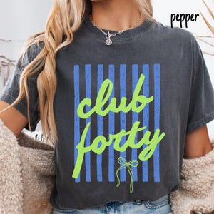 May include: A charcoal gray short-sleeve t-shirt featuring a graphic design with vertical blue stripes and the bright lime green text "club forty" written in a cursive script. A small lime green line-drawn bow is positioned below the text.