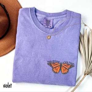 May include: A violet-colored t-shirt with a crew neck and short sleeves. The shirt features a butterfly graphic with the text "busy growing a tiny human." A gold necklace with a pendant is draped across the shirt.