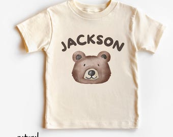 May include: A cream-colored t-shirt featuring a brown bear face graphic with the name "JACKSON" above it in brown letters. The word "natural" is printed in black at the bottom of the shirt. The shirt is made of soft material.