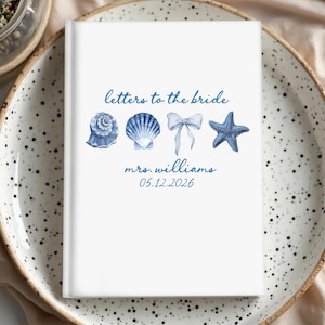 May include: White journal with blue watercolor illustrations of seashells, a bow, and a starfish. The text "letters to the bride" and "mrs. williams 05.12.2026" are written in blue script. The journal is on a speckled plate.