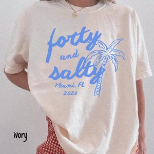 May include: Ivory-colored t-shirt with the text "forty and salty" in blue, along with a palm tree graphic. The text "Miami, FL 2026" is also printed on the shirt. The shirt is a casual, beach-themed design.