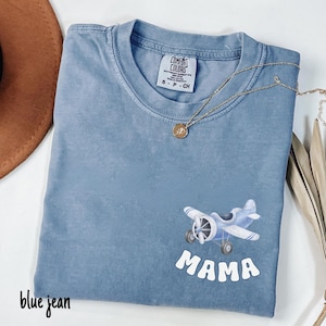 Pode incluir: Camiseta azul jeans com um pequeno gr&aacute;fico de avi&atilde;o e a palavra "MAMA" em branco. Um colar de ouro com um pingente repousa sobre a camisa. A camisa est&aacute; dobrada e tem uma etiqueta Comfort Colors.