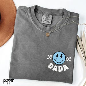 May include: A gray Comfort Colors t-shirt with a smiley face graphic and the word "DADA". The smiley face is blue with lightning bolt details and a checkered border. A silver necklace with a pendant is draped across the shirt.