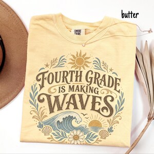 May include: A butter yellow short-sleeve t-shirt featuring a graphic print with the text "FOURTH GRADE IS MAKING WAVES". The design includes a sun, ocean waves, seashells, and a flower in shades of brown, blue, and cream, with a Comfort Colors label.