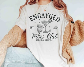 May include: White t-shirt with the text "Engayged Est 2024 Wives Club Sarah & Melissa" and a drawing of two cocktails.