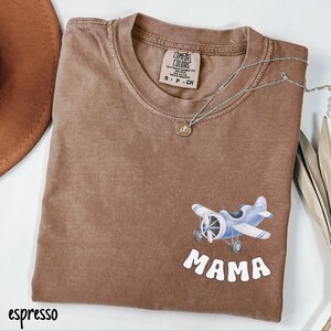 Pode incluir: Camiseta cor de caf&eacute; com um gr&aacute;fico de avi&atilde;o azul e a palavra "MAMA" em branco. A camiseta tem gola redonda e mangas curtas. Um colar de prata com um pequeno pingente est&aacute; drapeado sobre a camiseta.