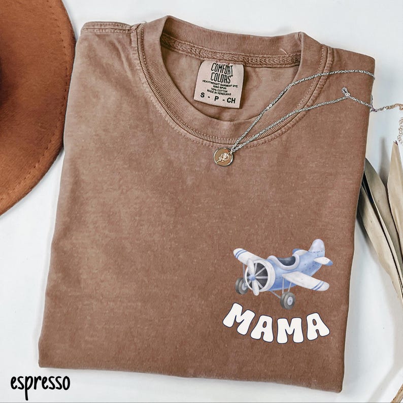 Pode incluir: Camiseta cor de caf&eacute; com um gr&aacute;fico de avi&atilde;o azul e a palavra "MAMA" em branco. A camiseta tem gola redonda e mangas curtas. Um colar de prata com um pequeno pingente est&aacute; drapeado sobre a camiseta.