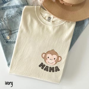 May include: Ivory-colored t-shirt with a brown monkey graphic and the word "MAMA" in brown letters. The shirt is made of a soft material and has a crew neck. A tan hat and blue jeans are in the background.