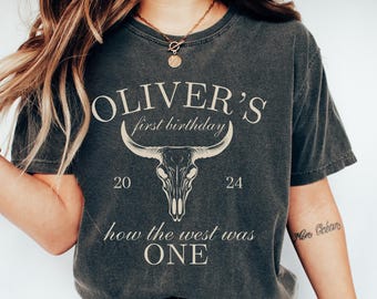 May include: A dark gray t-shirt with a white graphic design. The graphic features a skull with horns, the text "Oliver's first birthday", and the numbers "20" and "24". The text "how the west was one" is also included in the design.