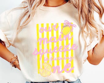 May include: An ivory t-shirt featuring a graphic of yellow vertical stripes, a lemon with a pink bow, and a lemon slice. The text reads, "our main squeeze is turning one" in pink cursive lettering. The shirt is paired with blue denim jeans.