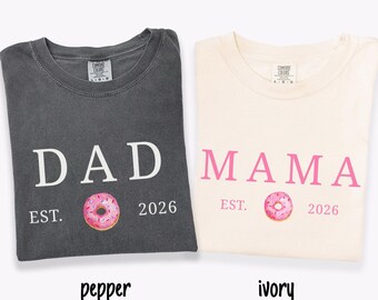 May include: Two t-shirts, one pepper-colored and one ivory-colored, are displayed. The pepper shirt says "DAD" with a pink donut graphic and "EST. 2026". The ivory shirt says "MAMA" with a pink donut graphic and "EST. 2026".
