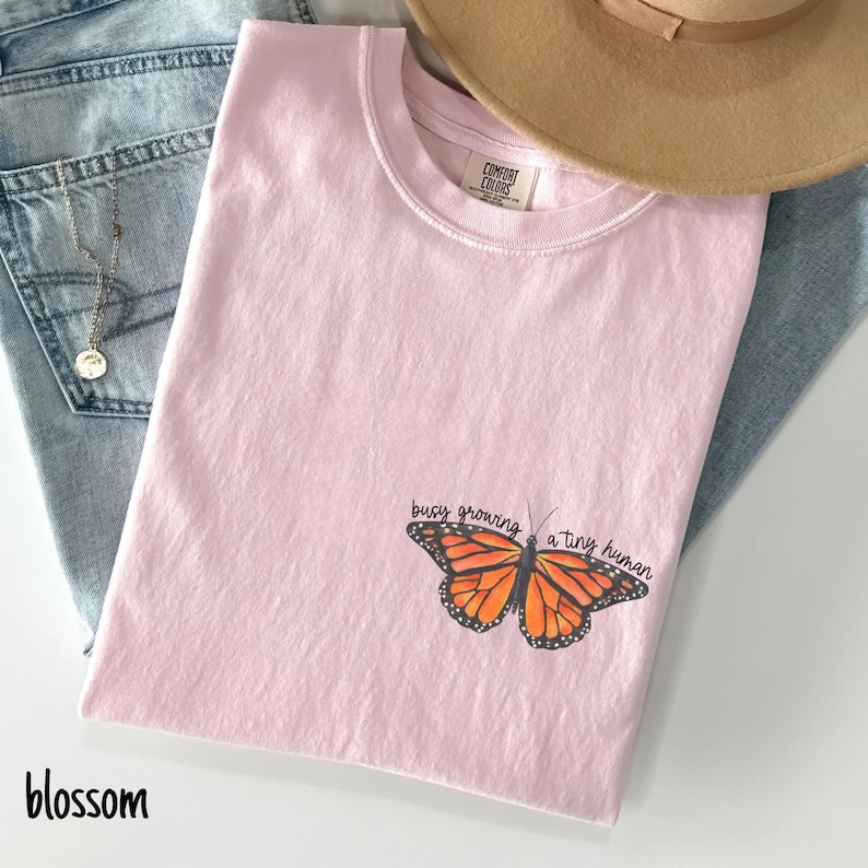 May include: A light pink t-shirt with a butterfly graphic and the text "busy growing a tiny human." The butterfly is orange and black. A tan hat and blue jeans are in the background.