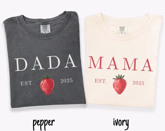 May include: Two t-shirts, one pepper-colored and one ivory-colored, are displayed. The pepper shirt says "DADA" with "EST. 2025" and a strawberry graphic. The ivory shirt says "MAMA" with "EST. 2025" and a strawberry graphic.