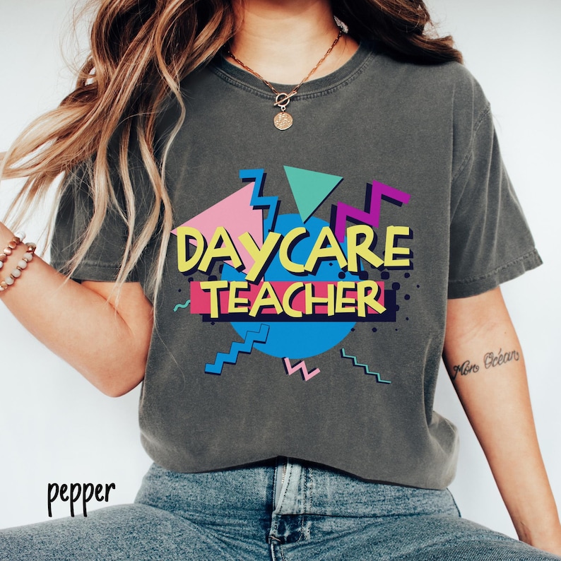 May include: Dark gray t-shirt with the words "DAYCARE TEACHER" in yellow, pink, blue, and purple, with a retro 80s design. The shirt has a round neck and short sleeves.