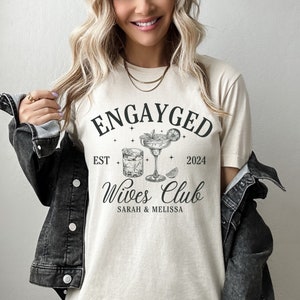 May include: A beige t-shirt with the text "Engayged Est 2024 Wives Club Sarah & Melissa" and a graphic of two cocktail glasses with a lime wedge.
