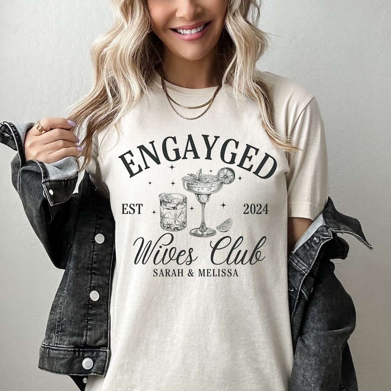 May include: A beige t-shirt with the text "Engayged Est 2024 Wives Club Sarah & Melissa" and a graphic of two cocktail glasses with a lime wedge.