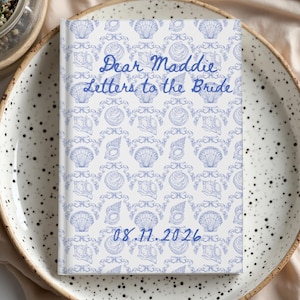 May include: A white journal with a blue seashell pattern and the text "Dear Maddie Letters to the Bride" and the date "08.11.2026". The journal is on a speckled white plate.