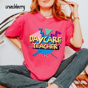 May include: A bright pink t-shirt with the words "DAYCARE TEACHER" in a retro, colorful design. The design includes geometric shapes in teal, purple, and yellow. The brand name "crunchberry" is in the top left corner.