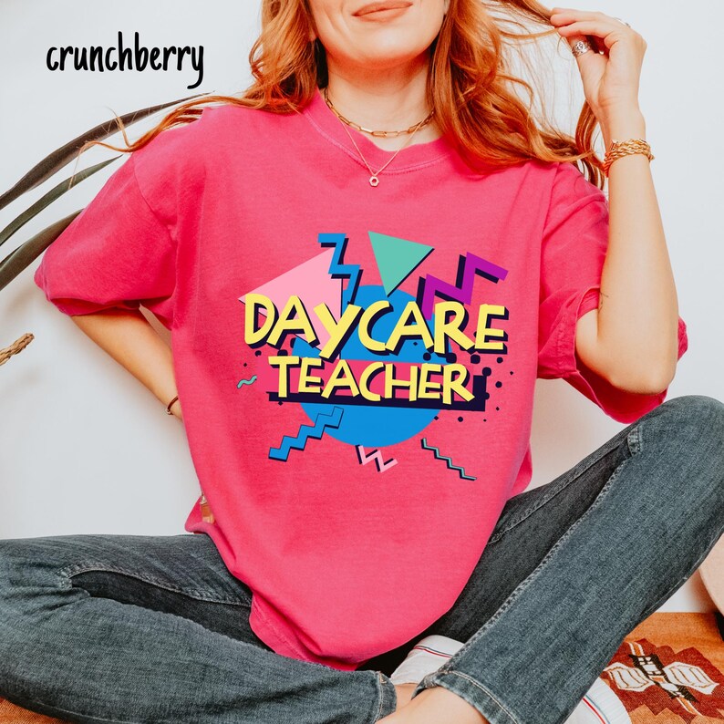 May include: A bright pink t-shirt with the words "DAYCARE TEACHER" in a retro, colorful design. The design includes geometric shapes in teal, purple, and yellow. The brand name "crunchberry" is in the top left corner.