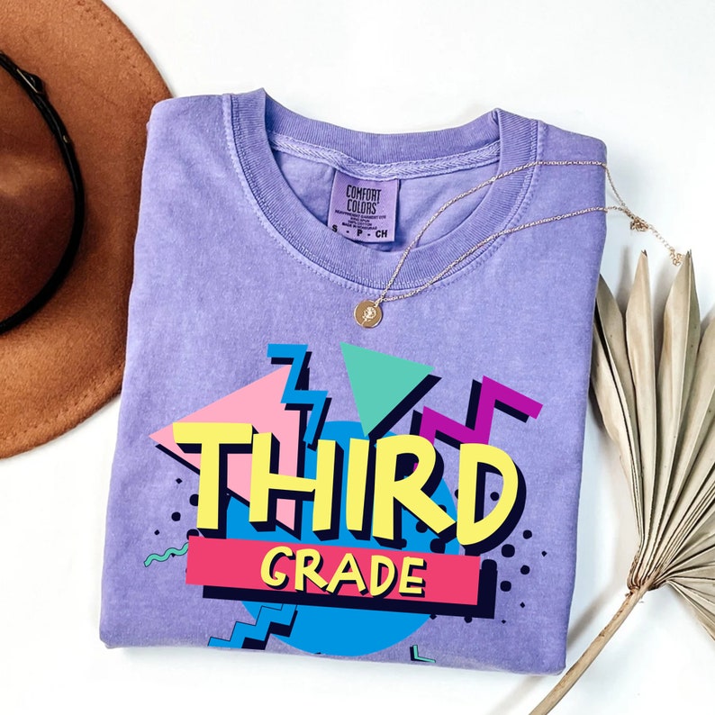 Op de afbeelding: Een lavendelkleurig Comfort Colors T-shirt met een retro graphic met de tekst "THIRD GRADE" in vette, kleurrijke letters. Het ontwerp bevat geometrische vormen in roze, blauw, geel en turquoise. Een gouden ketting en een bruine hoed zijn ook zichtbaar.