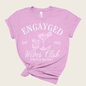 May include: Pink t-shirt with the text "Engayged Est 2024 Wives Club Sarah & Melissa" in white. The design also includes a cocktail glass and a glass of whiskey.