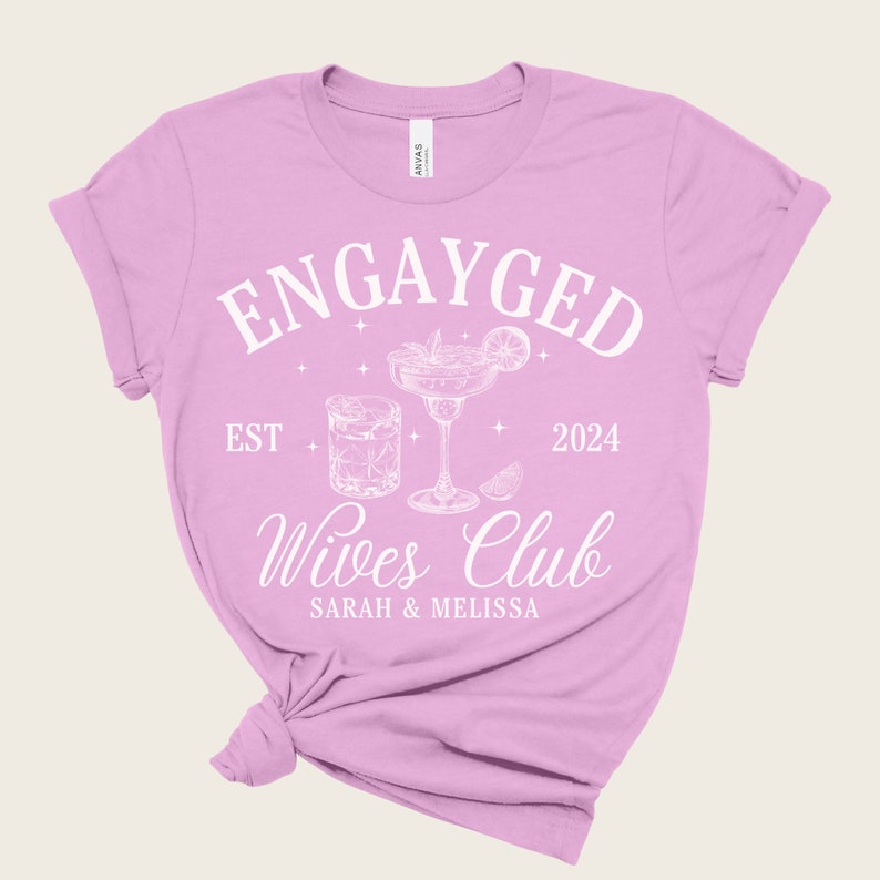 May include: Pink t-shirt with the text "Engayged Est 2024 Wives Club Sarah & Melissa" in white. The design also includes a cocktail glass and a glass of whiskey.