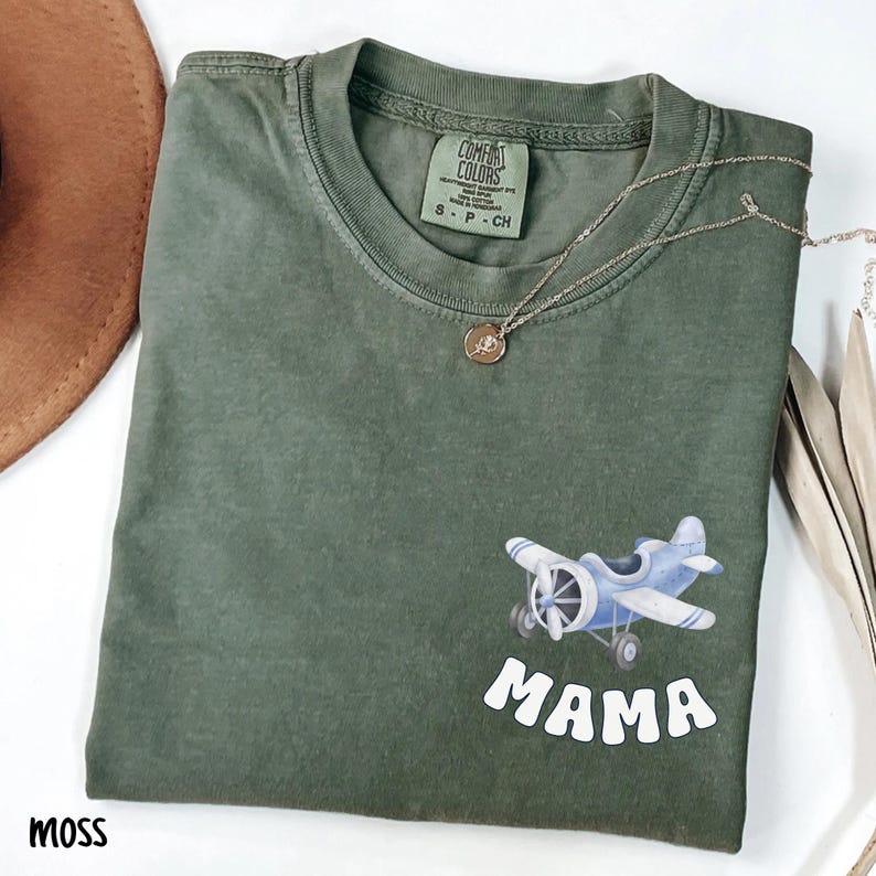 Pode incluir: Camiseta Comfort Colors verde musgo com um gr&aacute;fico de avi&atilde;o azul claro e a palavra "MAMA" em branco. Um colar de prata com um pequeno pingente est&aacute; drapeado sobre a camisa. A camisa &eacute; dobrada e tem um ajuste descontra&iacute;do.