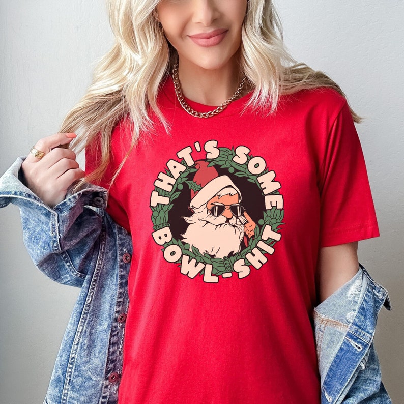 May include: Red t-shirt featuring a graphic of Santa Claus wearing sunglasses and a wreath that reads "That's Some Bowl-Shit".