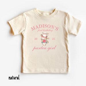 May include: A natural colored t-shirt with a pink graphic that says "MADISON'S first birthday 20 partea girl 25" with a stack of teacups in the center.