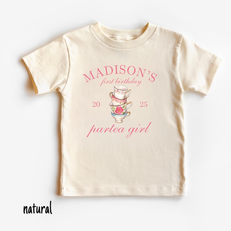 May include: A natural colored t-shirt with a pink graphic that says "MADISON'S first birthday 20 partea girl 25" with a stack of teacups in the center.